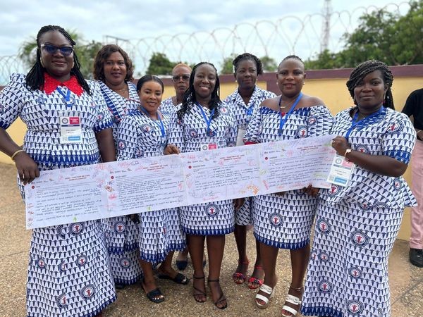 A group of eight women pose for a photo, holding a large, unraveled sheet of paper and smiling together. UNIGlobal Union.